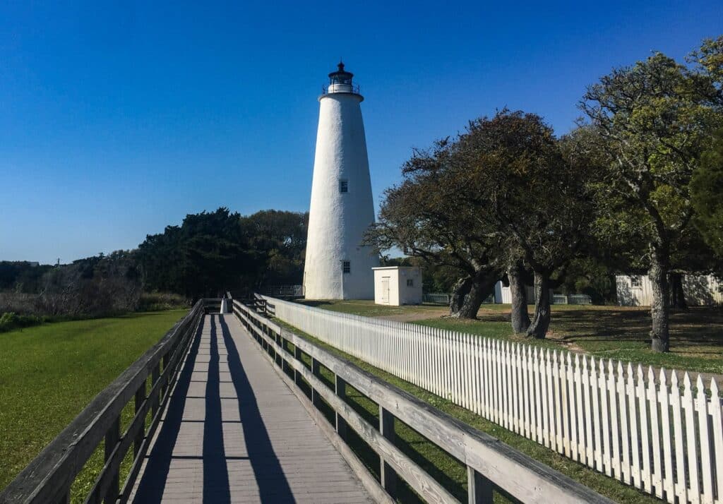 A long white-railed boardwalk leading through trees toward the Ocracoke Lighthouse.