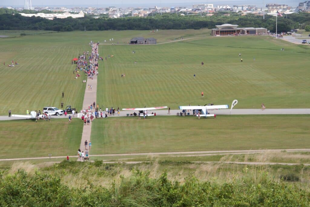 Small aircraft parked on grass beside the First Flight airstrip with the memorial monument on the hill beyond.
