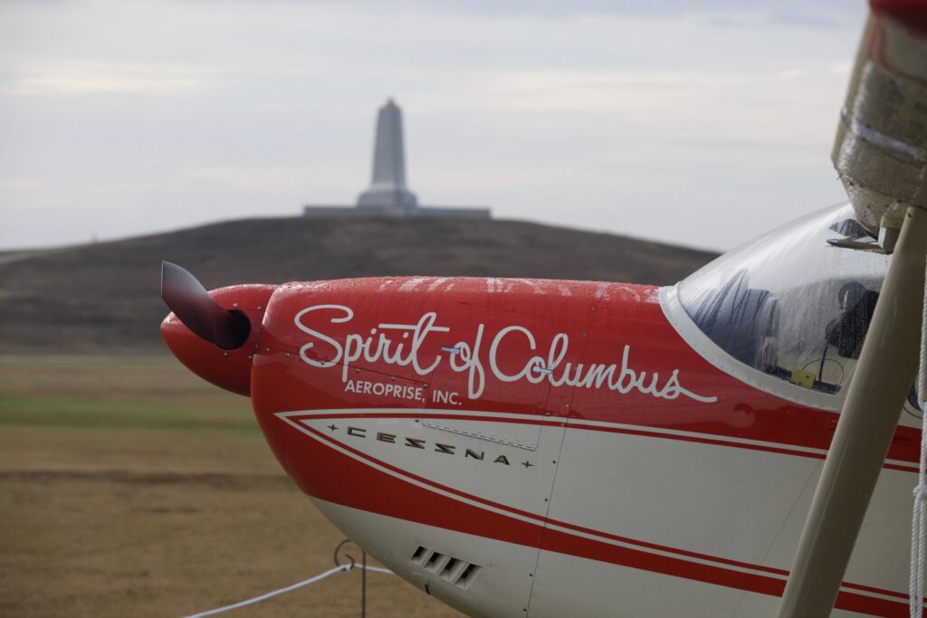 A vintage single-engine aircraft on the grass with the Wright Brothers Memorial monument on the hill behind it.