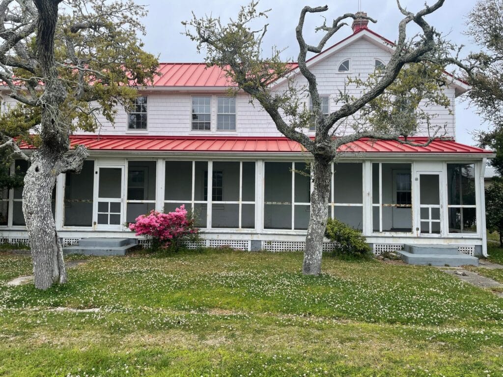 The white clapboard Double Keepers' Quarters at the Ocracoke Light Station, with a red roof and flowering shrubs.